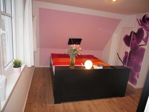 Una habitación con un mostrador de recepción negro, una pared rosa, una ventana con cortinas blancas, una pequeña planta en maceta y un televisor montado en la pared.