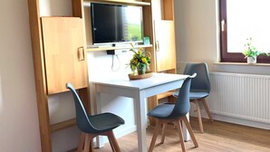 A room with a white table, two chairs, a television on a wooden shelf, a window with a view of greenery, and a radiator. - Groot Huus Horumersiel OG rear - Groot Huus upper floor for max. 4 persons (Wangerland)