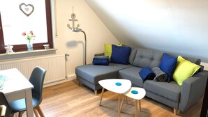 A cozy living room with a grey sofa, a small white table, and a window with a heart decoration. - Groot Huus Horumersiel OG rear - Groot Huus upper floor for max. 4 persons (Wangerland)