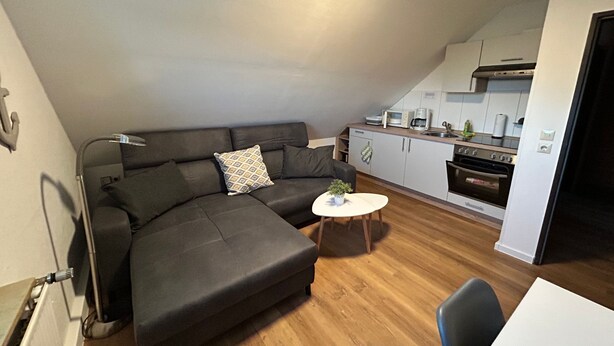 A modern living area with a sectional sofa, a small round table, and a kitchen in the background. - Groot Huus Horumersiel OG rear - Groot Huus upper floor for max. 4 persons (Wangerland)