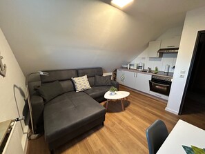A modern living area with a sectional sofa, a small round table, and a kitchen in the background. - Groot Huus Horumersiel OG rear - Groot Huus upper floor for max. 4 persons (Wangerland)