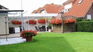 A residential backyard with a covered patio, a lawn, and flower beds. - Groot Huus Horumersiel OG rear - Groot Huus upper floor for max. 4 persons (Wangerland)