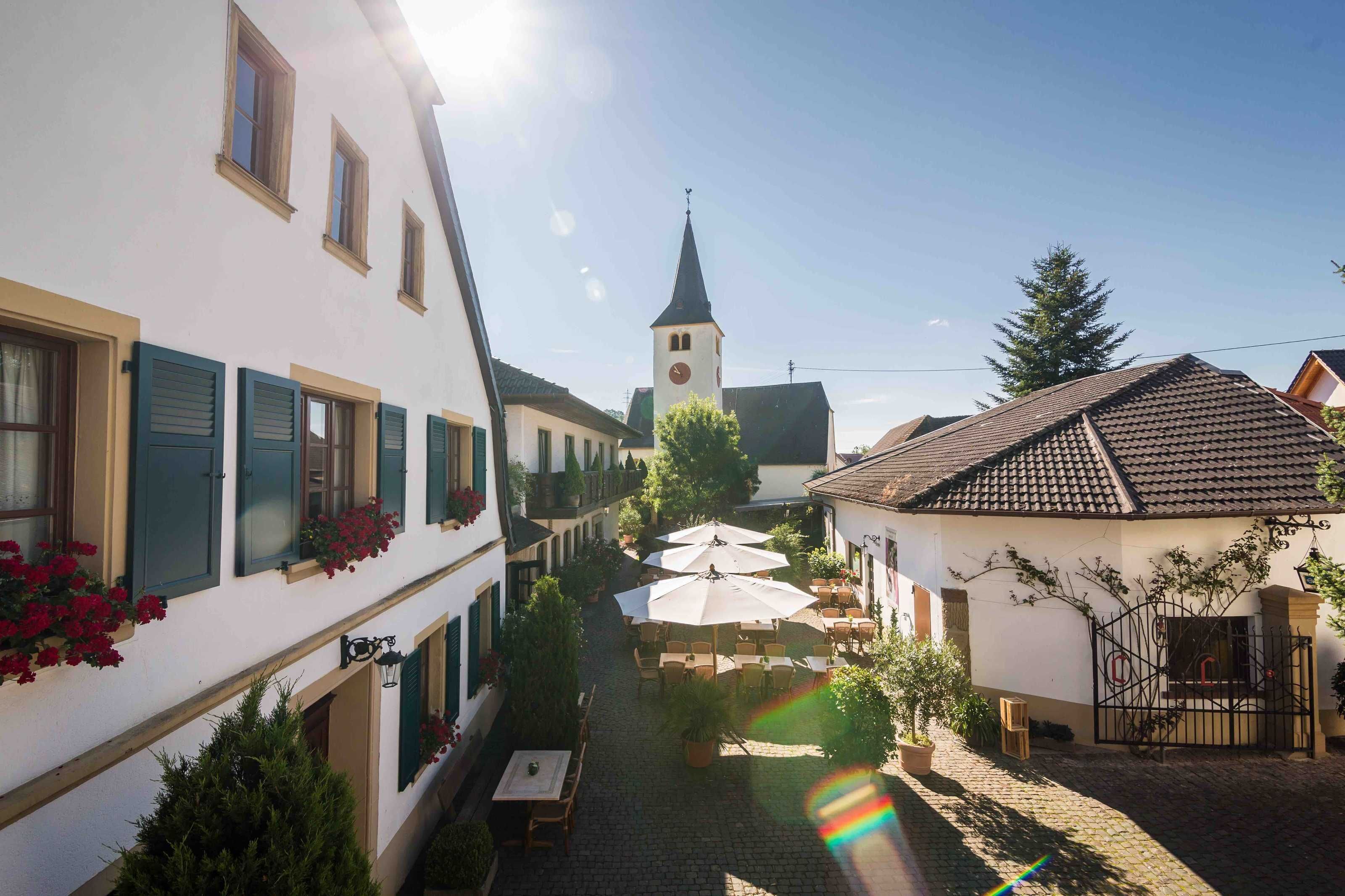 Une rue étroite bordée de bâtiments des deux côtés, un clocher d’église, des tables et chaises installées à l’extérieur sous des parasols, et un chemin pavé.