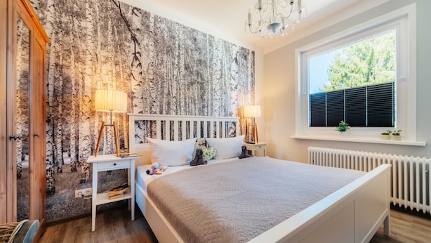 Een slaapkamer met een groot bed, een wit hoofdeinde, en een wand met een boomschorspatroon. Er staan twee nachtkastjes met lampen, een raam met zonwering, en een radiator.