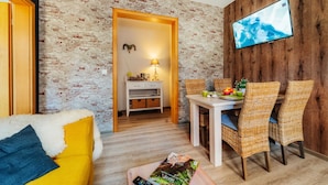 Een woonkamer met een gele bank, een eettafel met rieten stoelen, een aan een houten muur bevestigde televisie en een muur met een baksteenmotief.