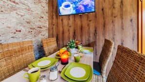 Een eethoek met een houten wand, een televisie boven de tafel, rieten stoelen, en een tafel gedekt met groene borden, bekers en een kaars.