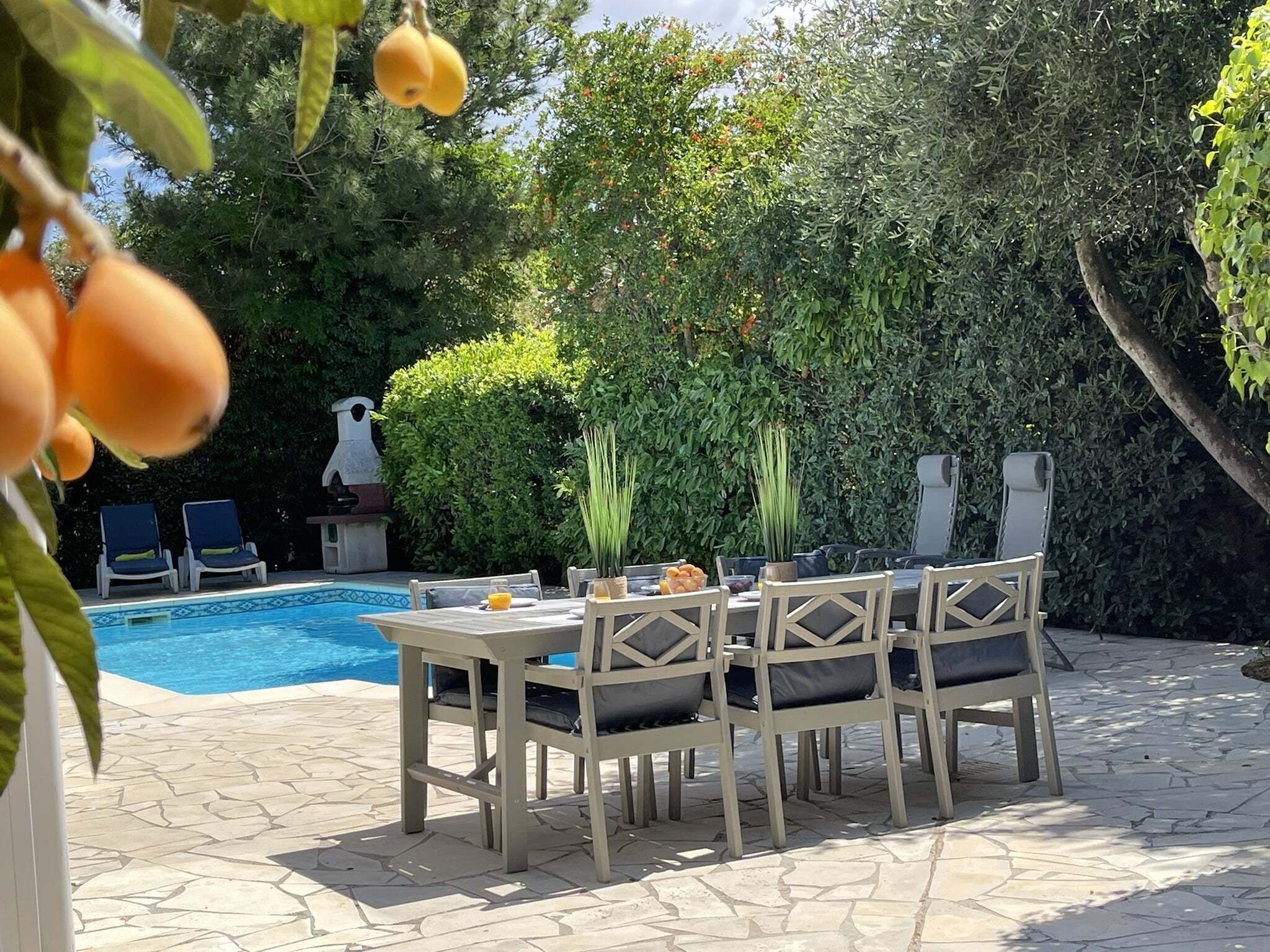 A poolside area with a table and chairs, a pool, and a hedge in the background.
