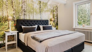 A bedroom with a large bed, two bedside tables, a window with a view of trees, and a wall with a forest wallpaper. - Bergwald - Ferienwohnung Bergwald (Braunlage)