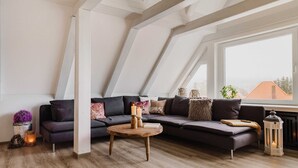 A modern loft apartment with a large window, a dark-colored sectional sofa, a wooden coffee table, and a decorative lantern. - Bergwald - Ferienwohnung Bergwald (Braunlage)