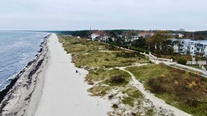 A coastal beach with a sandy shore, a grassy embankment, and residential buildings in the background. - Arkona Beach, Deutschland (Breege)