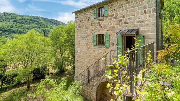 Un bâtiment en pierre avec deux fenêtres et une porte, entouré d’une végétation luxuriante et avec une colline en arrière-plan.