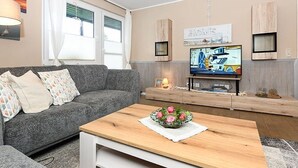 A living room with a grey sofa, a wooden coffee table with a floral centerpiece, a television on a wooden stand, and a wall-mounted shelf with decorative items. - Ferienwohnung Haus Heimathafen (Esens)