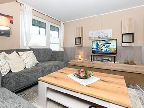A living room with a grey sofa, a wooden coffee table with a floral centerpiece, a television on a wooden stand, and a wall-mounted shelf with decorative items.