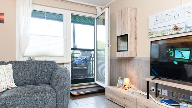 A living room with a grey sofa, a TV on a wooden stand, and a sliding glass door leading to an outdoor area. - Ferienwohnung Haus Heimathafen (Esens)