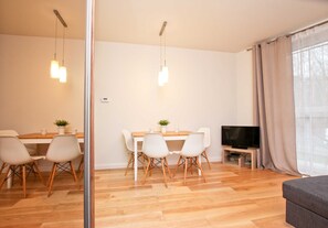 A modern living room with a wooden floor, a dining area with a white table and chairs, a flat-screen TV, and a large window with curtains. - Bałtycka (BA10) - BA10 (Swinoujscie)