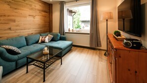 Ein Wohnzimmer mit einem türkisfarbenen Sofa, einem Couchtisch, einem Holzschrank, einem Fernseher und einem Fenster mit Vorhängen.