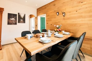 Ein Essbereich mit einem Holztisch, gedeckt für vier Personen, umgeben von schwarzen Stühlen. An der Wand hängen zwei gerahmte Bilder, ein Holzpaneel mit Geweihen und im Hintergrund eine grüne Tür.