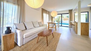 Una sala de estar moderna con un sofá, una mesa de centro de madera y una zona de comedor con una mesa y sillas. Hay un ventanal grande que proporciona luz natural y vistas al exterior.