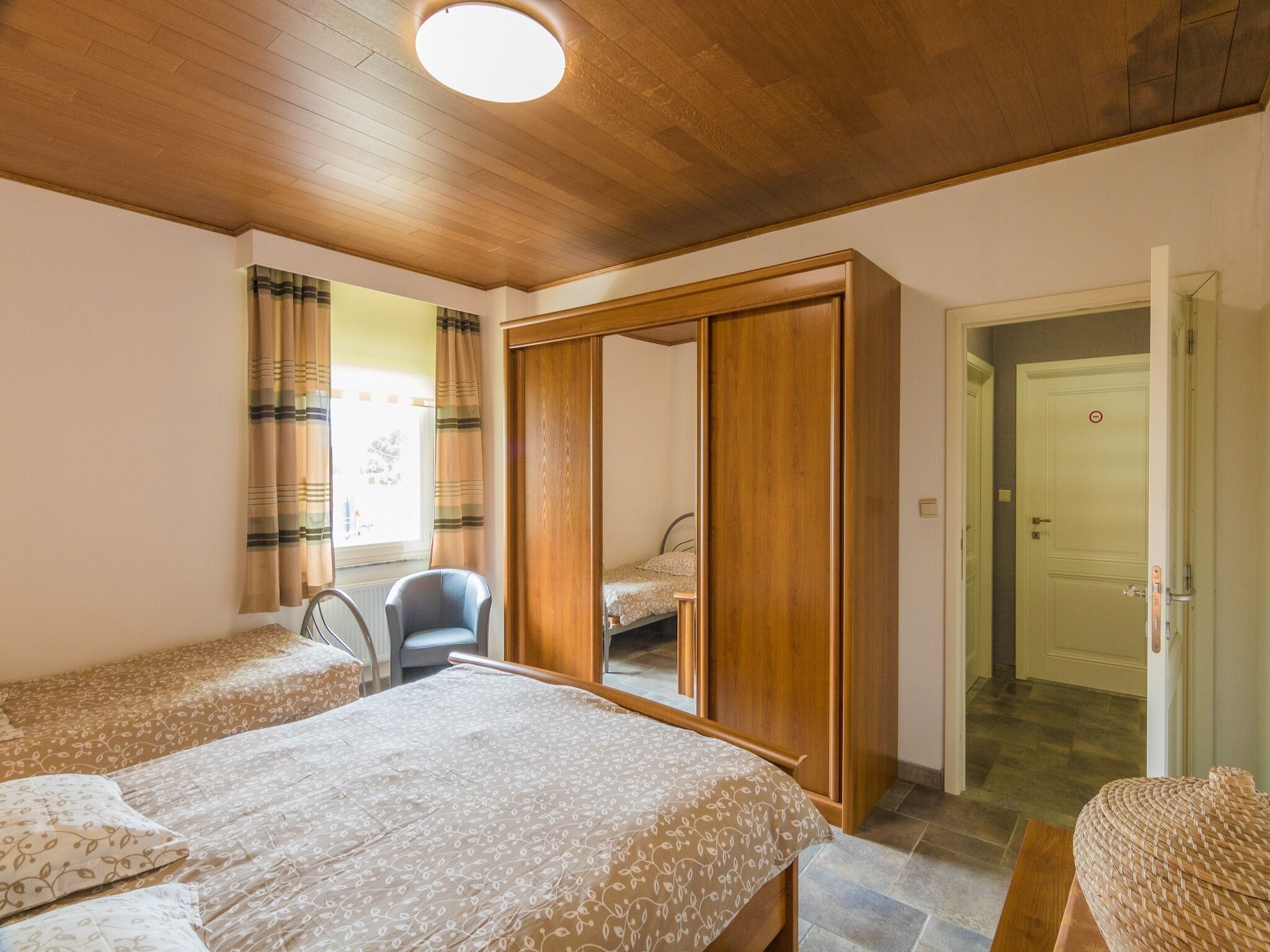A bedroom with two beds, a wooden wardrobe, a chair, and a door leading to another room.