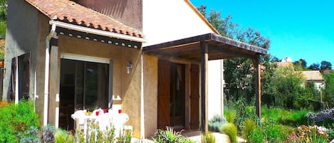 A small house with a tiled roof, a covered porch, and a garden with various plants and flowers.