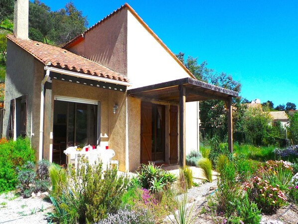 A small house with a tiled roof, a covered porch, and a garden with various plants and flowers.