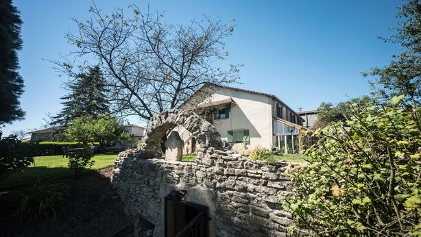 A stone structure with a wooden door, surrounded by greenery and a modern building in the background.