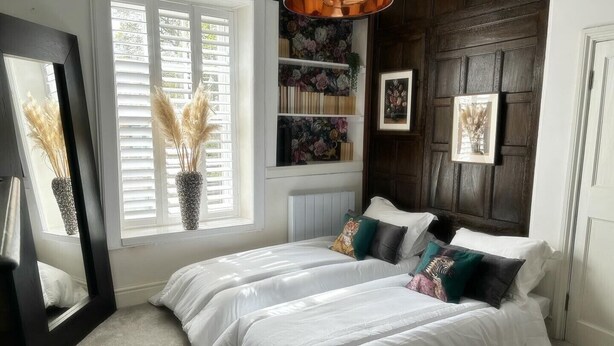 Une chambre avec un grand lit, une fenêtre avec des stores blancs, un miroir, une bibliothèque remplie de livres et d’objets décoratifs, et une armoire en bois sombre.