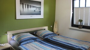 Um quarto com cama, mesa de cabeceira, luminária, quadro na parede, janela com persianas e planta em vaso.