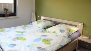 A bed with a leaf-patterned bedspread, a bedside table with a lamp, and a window with a plant. - Ferienwohnung 6 - Ferienhaus Luise Urlaubslust Trassenheide (Trassenheide)