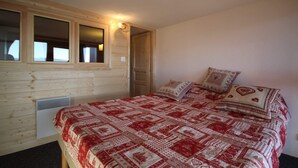 A bedroom with a wooden exterior wall, a window, a bed with patterned bedding, and a wall-mounted light fixture. - Arcs 1800, Vogel building, Mont-Blanc view, 200m from slopes and shops (Bourg-Saint-Maurice)