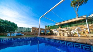 A swimming pool with a stone border, outdoor seating, and a covered area with a table and chairs.