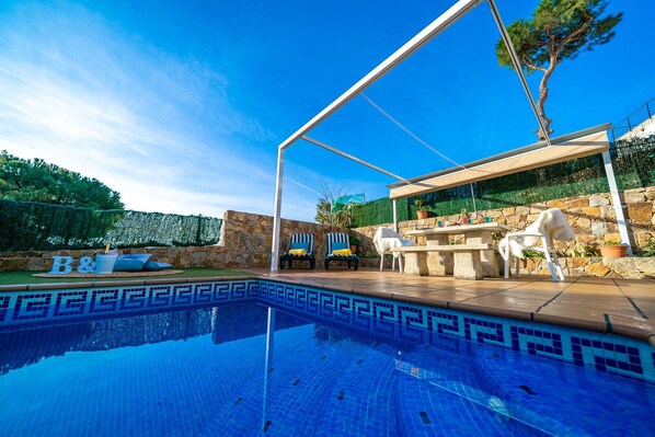 A swimming pool with a stone border, outdoor seating, and a covered area with a table and chairs.