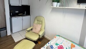 A small, modern living space with a kitchenette, a green armchair, a sofa with a colorful blanket, and a potted plant on a shelf.