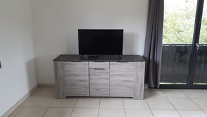 A modern, minimalist room with a grey cabinet featuring a flat-screen TV on top, a sliding glass door to a balcony, and a tiled floor.
