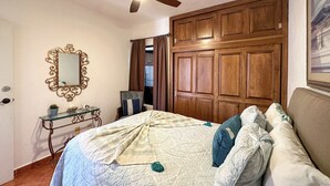 Quarto com cama, ventoinha no teto, guarda-roupa de madeira, mesa de consola de vidro com espelho e um quadro na parede.