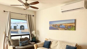 Sala de estar com sofá, poltrona e uma mesinha com uma planta. Há um ventilador de teto, um ar-condicionado e uma janela com cortinas.