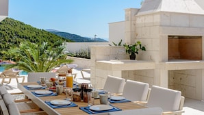 An outdoor dining area with a stone barbecue, white furniture, and a view of a pool and hills. - Cozy home in Marina with sauna (Marina)