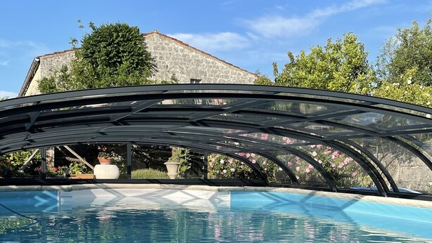 Una piscina con cerramiento de vidrio, rodeada de follaje y un edificio de piedra al fondo.