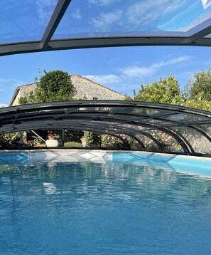 Una piscina con cerramiento de vidrio, rodeada de follaje y un edificio de piedra al fondo.