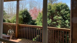 A wooden deck with a railing, a table, and chairs, overlooking a forested area with trees and a clear sky.