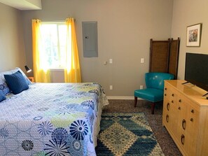 Une chambre à coucher avec un lit, une commode, une chaise, un téléviseur et une fenêtre avec des rideaux.