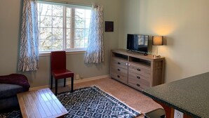 A small, well-lit room with a window, a television, a wooden cabinet, a red chair, and a patterned rug.