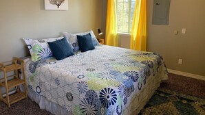 A bedroom with a bed, a nightstand, a wall-mounted shelf, a window with yellow curtains, and a power outlet.