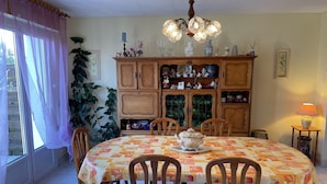 A dining room with a round table covered in a patterned tablecloth, surrounded by wooden chairs. There is a chandelier above the table, and a cabinet with glass doors and various items on top. - lovely quiet house (Lanester)