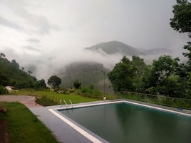 Una piscina rectangular con un paseo perimetral, ubicada en un paisaje verde y exuberante con árboles y montañas brumosas al fondo.