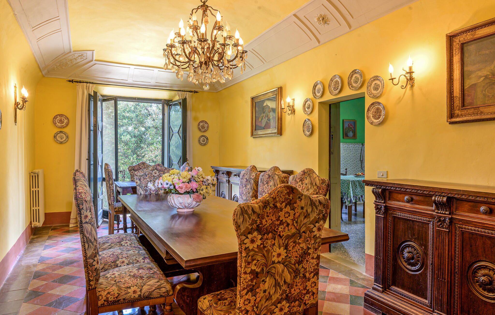 Ein Esszimmer mit langem Tisch, floralen Stühlen, einer Chandelieber und dekorativen Tellern an den Wänden.