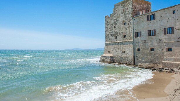 A coastal building with a tower and multiple windows, adjacent to a sandy beach and the sea.