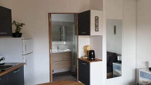 A compact hotel room with a kitchenette, a small bathroom, a bed, and a bedside table with a lamp. - BEAUTIFUL BRAND-NEW STUDIO APARTMENT (Le teich)