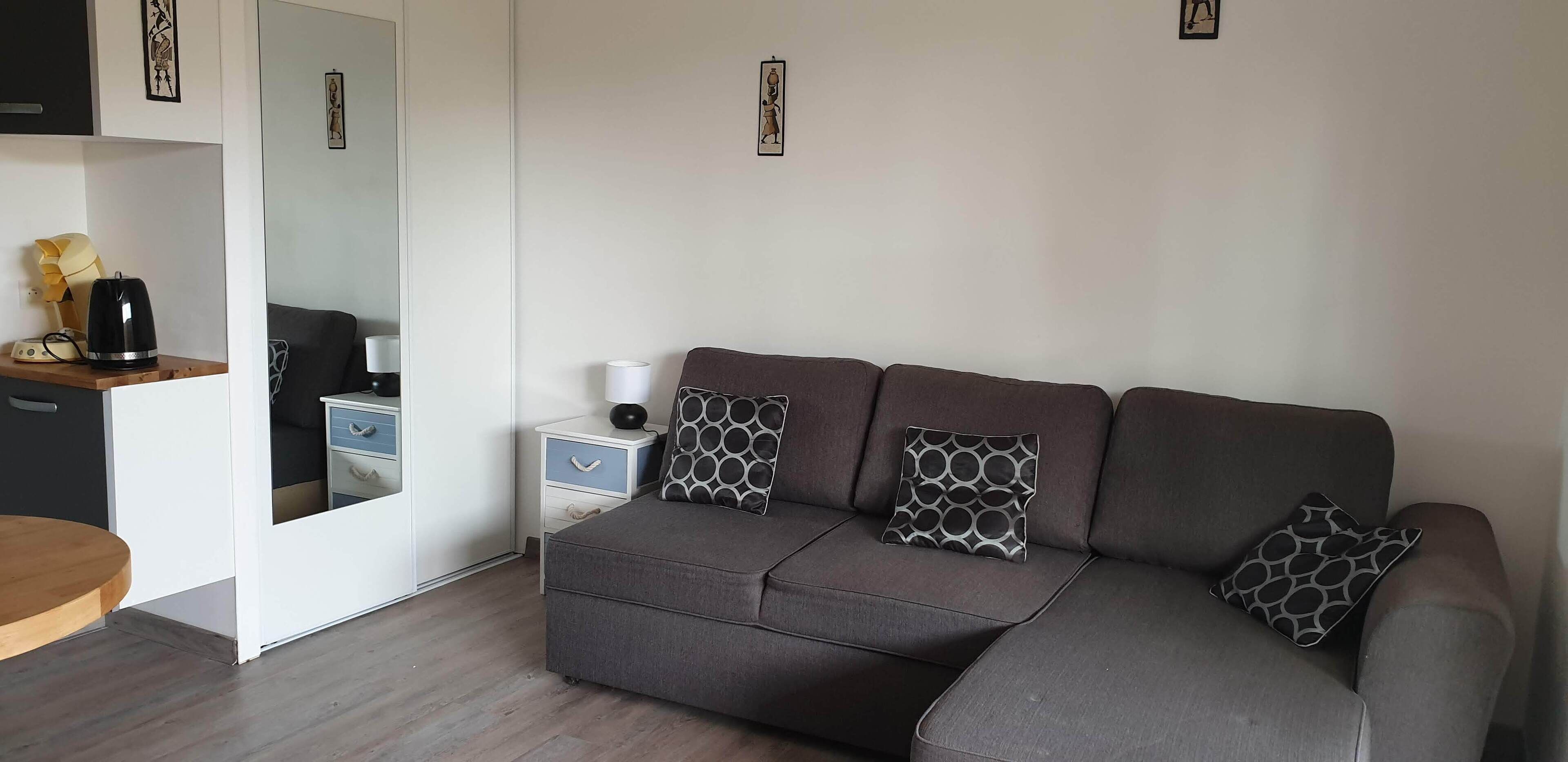 A modern living room with a grey sofa, patterned cushions, a side table with a lamp, and a coffee maker on a wooden shelf.