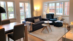 Een moderne woonkamer met een bank, stoelen, een salontafel en een eethoek. Grote ramen bieden natuurlijk licht en uitzicht op een gebouw van rode baksteen.